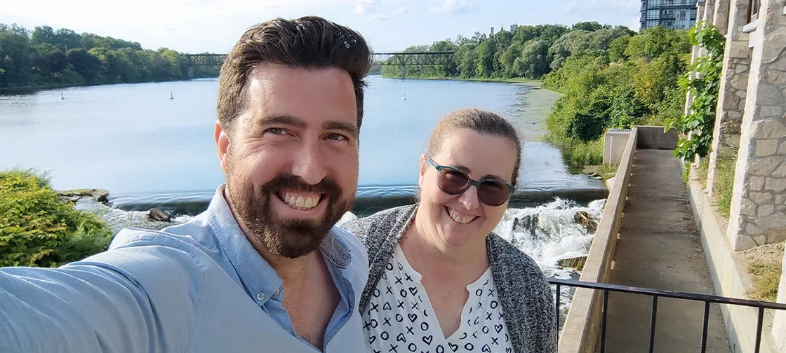 The same selfie without the Costco background: Jef and his mum on a walkway
                  above a river and weir, trees and a rail bridge behind them.